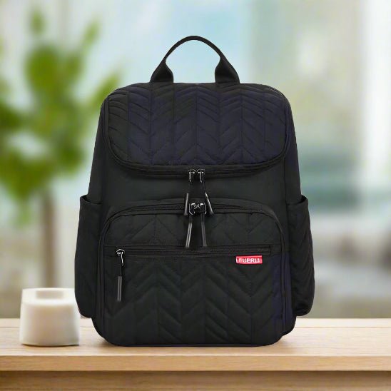 Le Sac Bebe à Langer - NappyBag a dos, noir et matelassé avec une fermeture à glissière sur le devant et des poches latérales, repose sur une table en bois à côté d'une petite tasse blanche. L'arrière-plan, légèrement flou, présente un feuillage vert et de la lumière.