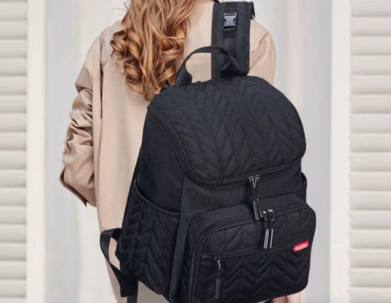 Une personne aux cheveux ondulés, châtain clair, vêtue d'un manteau beige, est vue de dos, portant un grand Sac Bebe à Langer - NappyBag a dos noir avec plusieurs compartiments à fermeture éclair et une petite étiquette rouge.