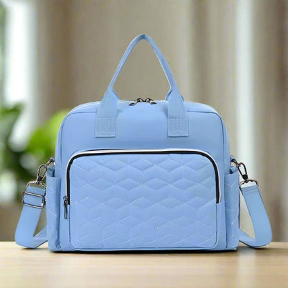 Le Sac bebe | Sac de Maternité, bleu clair avec motifs géométriques matelassés, grande capacité, deux anses supérieures, poche frontale zippée, poches latérales et bandoulière amovible est posé sur une surface en bois avec un arrière-plan flou.