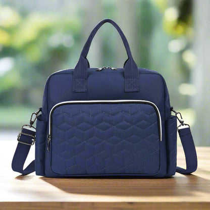 Le Sac bebe | Sac de Maternité est un sac à langer bleu marine avec une poche avant matelassée, une grande capacité, des poignées doubles, une bandoulière amovible et une fermeture à glissière, présenté sur une surface en bois avec un arrière-plan extérieur flou.