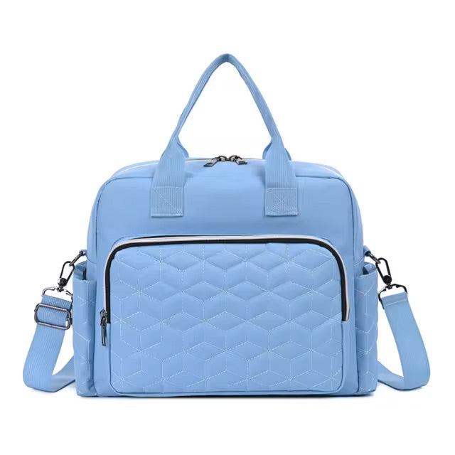 Le Sac bébé | Maternité est un sac à langer matelassé bleu clair avec deux anses courtes, une bandoulière amovible, une grande poche zippée sur le devant avec coutures hexagonales, une grande capacité, des tirettes de fermeture éclair en métal et des poches latérales.