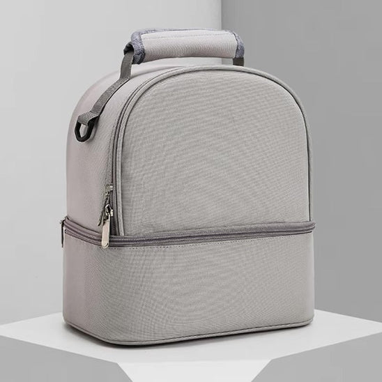 Le Sac Bebe - Petit est un sac rectangulaire gris clair avec deux compartiments, des fermetures à glissière, une poignée supérieure et un design minimaliste pour les performances thermiques, présenté sur une surface blanche avec un fond gris.
