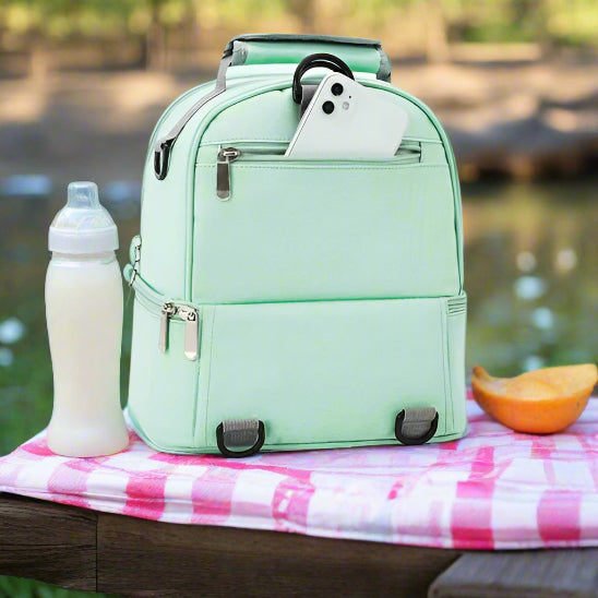 Le Sac Bébé - Petit vert clair est présenté sur un socle rond blanc. Il est doté de deux compartiments zippés et d'une poche avant avec un smartphone blanc partiellement visible.
