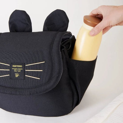 Une main place un biberon beige dans la poche latérale dun sac noir aux motifs doreilles de chat et de moustaches. Cet accessoire pratique pour tout Sac bébé - Poussette est doté dun rabat avec une petite étiquette affichant du texte.