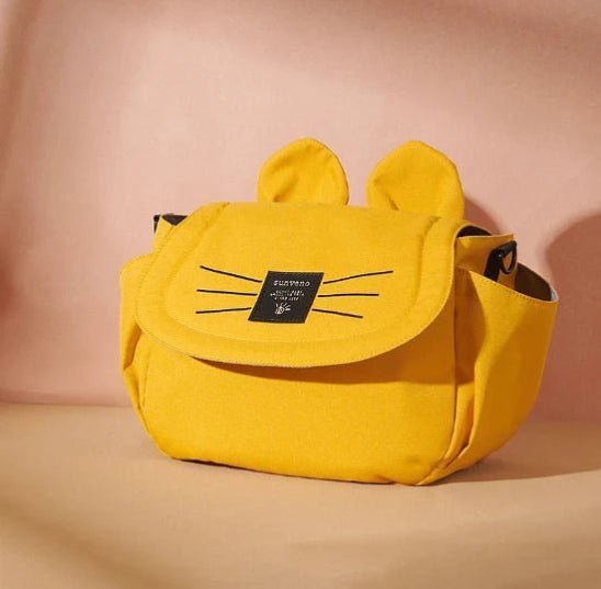 Le Sac bébé - Poussette, un sac jaune aux détails de moustaches noires et de petites oreilles, est présenté sur un fond beige. Conçu comme un accessoire pratique, il est doté dune fermeture à rabat et dune étiquette rectangulaire noire sur le devant. Idéal pour organiser les essentiels lors de vos déplacements.