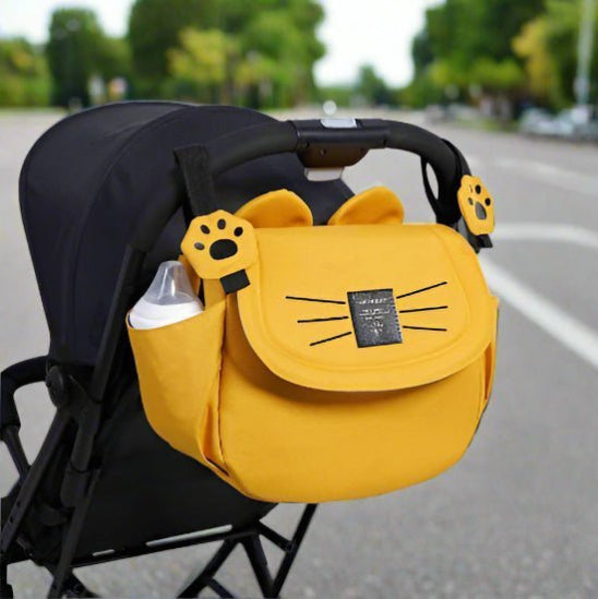 Le Sac bébé - Poussette est un sac de poussette en forme de tête de chat jaune avec des moustaches noires, des oreilles et des attaches en forme de pattes, attaché à une poignée de poussette. Il a une poche latérale montrant un biberon. Le décor est une rue bordée darbres.