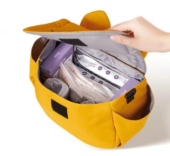 Une main ouvre le sac à langer jaune moutarde, laissant apparaître des couches, des lingettes pour bébé emballées en violet et un biberon à lintérieur. Le Sac bébé - Poussette offre plusieurs compartiments pour organiser les essentiels, ce qui en fait un accessoire pratique indispensable pour les parents en déplacement.