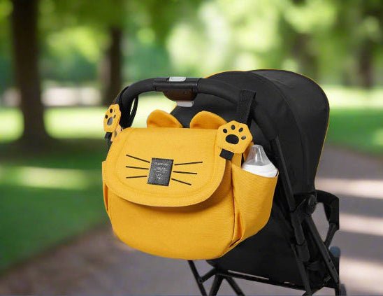 Le Sac bebe - Petit Sac Poussette, en tissu jaune avec des motifs de pattes et de moustaches de chat, s'attache à une poignée de poussette noire. Un biberon dans sa poche latérale rend cet organiseur idéal pour les promenades sur les chemins bordés d'arbres.