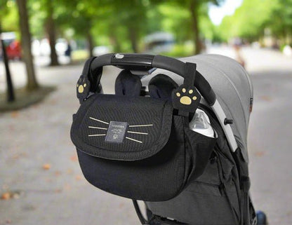 Le Sac bebe - Petit Sac Poussette, un organisateur noir avec un visage de chat, des moustaches et des pinces en forme de pattes, est attaché à la poignée d'une poussette grise sur un chemin bordé d'arbres, gardant vos objets essentiels organisés et facilement accessibles.
