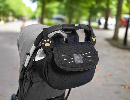 Le Sac bebe - Petit Sac Poussette, qui représente un visage de chat et des pinces à pattes, est attaché comme un accessoire ludique à la poignée d'une poussette grise sur un chemin bordé d'arbres.