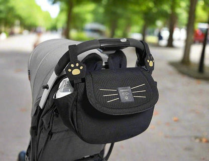 Le Sac bebe - Petit Sac Poussette, qui représente un visage de chat et des pinces à pattes, est attaché comme un accessoire ludique à la poignée d'une poussette grise sur un chemin bordé d'arbres.