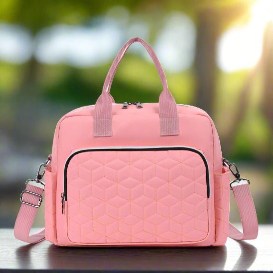 Le Sac bebe | Sac de Maternité est un sac à langer rose clair de grande capacité, doté d'une poche frontale hexagonale matelassée, de deux poignées courtes, d'une bandoulière amovible et d'une quincaillerie argentée, présenté sur une surface foncée avec un fond vert flou.
