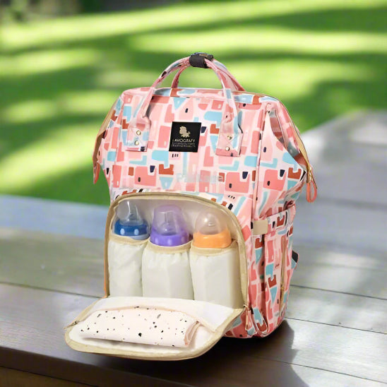 Un sac de maternité rose et multicolore aux motifs abstraits est présenté sur un fond rose. La poche avant est ouverte, révélant trois biberons dans des poches séparées et un tissu plié.