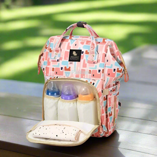 Un sac de maternité rose et multicolore aux motifs abstraits est présenté sur un fond rose. La poche avant est ouverte, révélant trois biberons dans des poches séparées et un tissu plié.