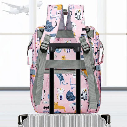 Un Sac pour Bébé | Chat, comportant des illustrations colorées de chats et un système d'organisation intelligent, est attaché à une poignée de valise argentée, tandis qu'un avion vole à l'extérieur des grandes fenêtres en arrière-plan.