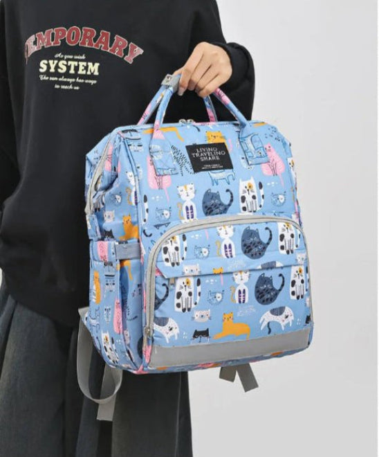 Une personne portant un sweat-shirt noir tient le sac à dos bleu « Sac pour Bebe | Chat », orné d'un motif de chat coloré. Ce sac polyvalent offre un système d'organisation intelligent avec de multiples poches et fermetures éclair pour garder tout en place.