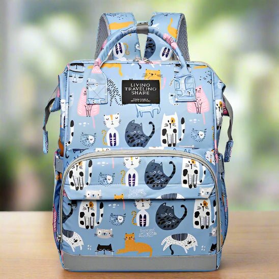 Un Sac pour Bebe | DesignChat bleu clair avec des imprimés de chats colorés et une étiquette LIVING TRAVELING SHARE sur la poche avant, présenté sur une surface en bois sur un fond vert flou.
