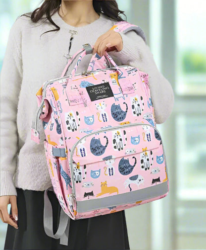 Une personne portant un pull gris tient le Sac pour Bebe | Chat, un sac à dos rose orné d'illustrations de chats de dessins animés. Doté de multiples poches et d'un système d'organisation intelligent, il est présenté sur un fond blanc uni avec la personne partiellement visible.