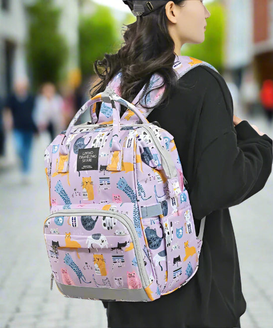 Une personne aux cheveux longs, vêtue d'une chemise noire et d'une casquette, porte un sac à dos violet « Sac pour Bébé | Chat » orné de motifs de chats ludiques et de la phrase « VIVRE VOYAGER PARTAGER », soulignant son utilisation polyvalente comme sac polyvalent pour toute aventure.