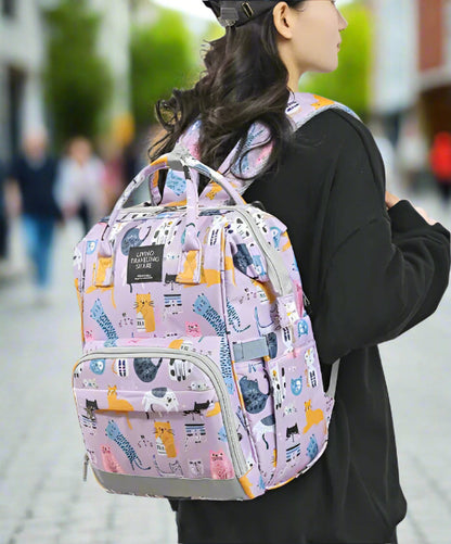 Une personne aux cheveux longs, vêtue d'une chemise noire et d'une casquette, porte un sac à dos violet « Sac pour Bébé | Chat » orné de motifs de chats ludiques et de la phrase « VIVRE VOYAGER PARTAGER », soulignant son utilisation polyvalente comme sac polyvalent pour toute aventure.