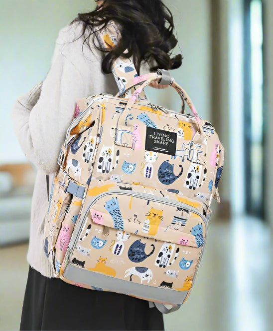 Une femme porte un sac à dos Sac pour Bebe | Chat avec des illustrations de chats et un système d'organisation intelligent, comprenant plusieurs compartiments, une poignée et des bretelles. Elle l'associe à un pull gris clair sur un fond blanc uni.