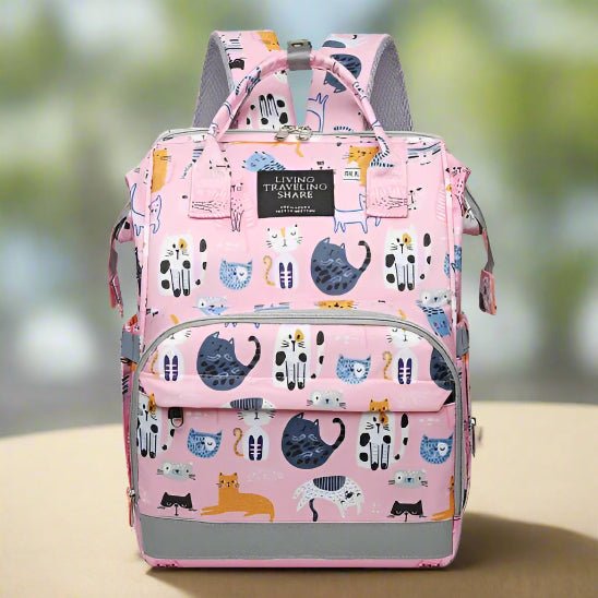 Le Sac pour Bebe | DesignChat est un sac à dos rose pour bébé avec des chats de dessins animés dans différentes poses et couleurs, plusieurs fermetures à glissière, des poches latérales et des bretelles réglables, sur un fond vert flou.