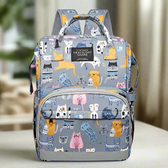Un Sac pour Bebe | DesignChat gris avec des illustrations de chats colorés, deux compartiments principaux, une poche frontale zippée et des bandoulières rembourrées est exposé à l'intérieur avec un arrière-plan flou.