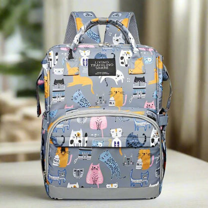 Un Sac pour Bebe | DesignChat gris avec des illustrations de chats colorés, deux compartiments principaux, une poche frontale zippée et des bandoulières rembourrées est exposé à l'intérieur avec un arrière-plan flou.
