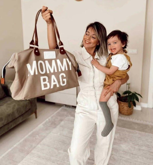 Une femme en combinaison blanche tient un grand sac beige pour bébé dans une main et son tout-petit souriant dans l'autre. Elles sont debout à l'intérieur, sur un tapis clair, près d'un canapé vert et d'une plante en pot, illustrant le style moderne de maman.