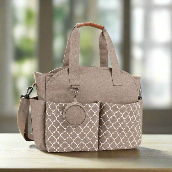 Le Sac pour Maternité - LeComplet pour bébé en beige et marron clair, avec des poches frontales à motifs, des poignées courtes, une bandoulière et un accessoire rond sur une poche, est posé sur une surface en bois avec une fenêtre floue à l'arrière.