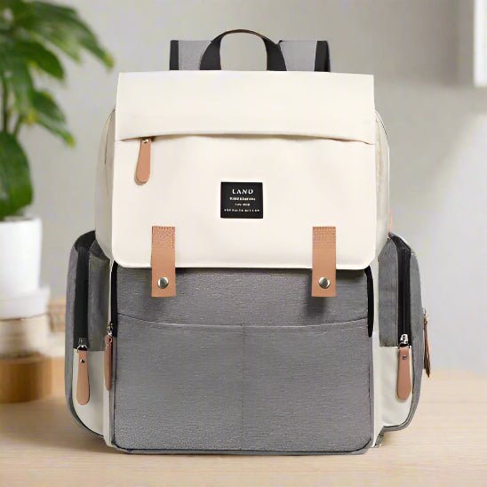 Le Sac pour la Maternité - Sac à dos présente un rabat beige chic, un corps gris, des accents de cuir marron, des poches latérales et une poignée noire - idéal pour les soins du bébé - présentés sur une surface claire avec une plante en arrière-plan.