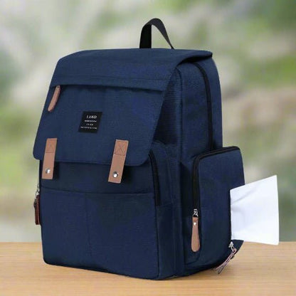 A Sac pour la Maternité - Sac à dos bleu foncé avec des accents marron repose sur du bois, un papier blanc visible dans la poche latérale zippée pour les soins de bébé. Le fond légèrement flou met en valeur son design élégant.