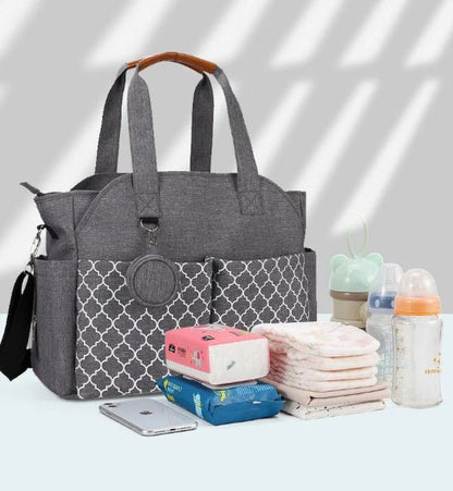 Un Sac pour Maternité - LeComplet pour bébé gris avec des poches frontales à motifs est placé à côté d'articles essentiels pour bébé - des lingettes humides, des couches, des biberons, un étui à sucette et un smartphone - disposés de manière ordonnée sur une surface claire.
