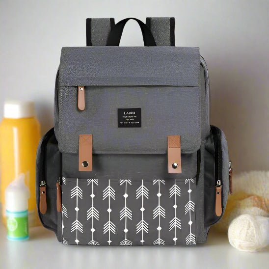 Le Sac pour la Maternité - Sac à dos est gris avec des bretelles marron, une poche avant imprimée d'une flèche blanche et une étiquette noire. Présenté à côté des articles de puériculture - biberons et serviettes - il est parfait pour les soins de bébé.