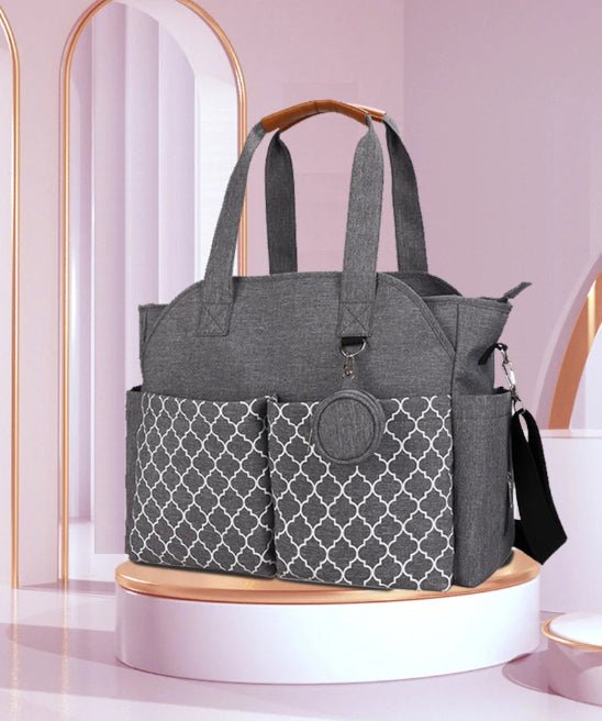 Le Sac pour Maternité - LeComplet pour bébé, de couleur grise avec des poches avant à motifs, repose sur un socle rond. Des arches incurvées, rehaussées d'or, et des murs rose pâle forment un arrière-plan doux et moderne.