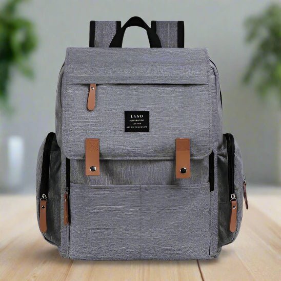 Le Sac pour la Maternité - Sac à dos est gris avec des accents de cuir marron, comporte de multiples poches frontales et latérales, une étiquette rectangulaire noire sur le devant, et est présenté sur une surface en bois avec un arrière-plan flou en intérieur.