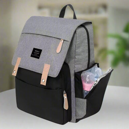 A Sac pour la Maternité - Sac à dos en gris et noir avec des accents tan est sur une surface blanche. Un biberon et un article transparent se trouvent dans la poche latérale, parfaits pour les soins de bébé. La poche principale est fermée ; une étiquette est apposée sur le rabat avant.