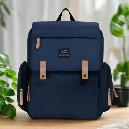 Le Sac pour la Maternité - Sac à dos, bleu marine avec des fermetures à glissière et des boucles de couleur havane, repose sur une surface en bois clair. Il comporte deux poches latérales et une étiquette frontale, idéale pour les soins de bébé. Des plantes vertes en pot décorent l'arrière-plan.