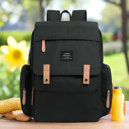 Le Sac pour la Maternité - Sac à dos en noir avec des fermetures à glissière de couleur havane et une poche à rabat sur le devant est présenté sur une table extérieure, à côté d'une bouteille jaune et d'un article en tricot, avec de la verdure et une grande fleur jaune dans l'arrière-plan flou.
