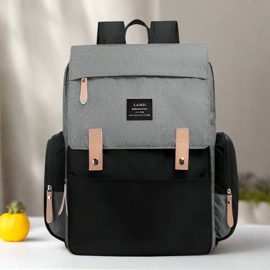 Le Sac pour la Maternité - Sac à dos, un élégant sac à dos bicolore avec un rabat gris, un corps noir, des tirettes tan, des poches latérales et une étiquette avec logo sur le devant, repose sur une surface blanche à côté d'un petit légume jaune.