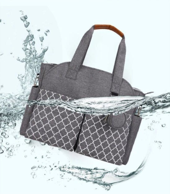 Un sac de maternité gris à motifs, d'une grande capacité et doté de deux poches avant, se présente sur un fond clair. Des éclaboussures d'eau le parcourent, soulignant sa polyvalence et sa résistance à l'eau.