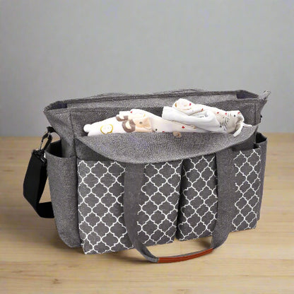Le Sac pour Maternité, gris à motif géométrique et grande capacité, repose sur du bois. Doté de multiples poches et poignées, il offre un accès facile à une couverture pour bébé à imprimé animal enroulée qui dépasse du compartiment principal.
