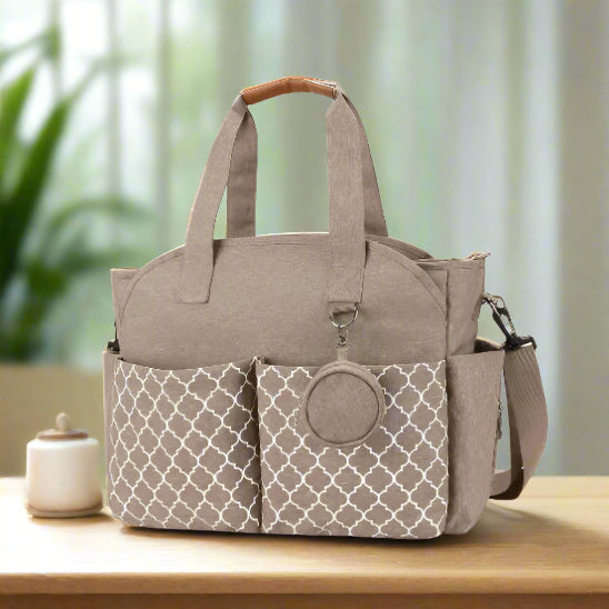 Le Sac pour Maternité est un sac en tissu beige de grande capacité, doté de deux poches avant à motifs, de deux poignées, d'une bandoulière amovible et d'une pochette ronde pour accessoires attachée sur le devant.