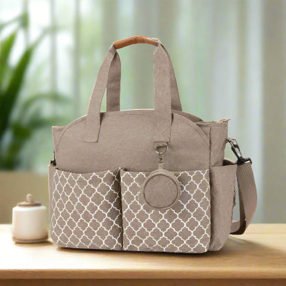 Le Sac pour Maternité est un sac en tissu beige de grande capacité, doté de deux poches avant à motifs, de deux poignées, d'une bandoulière amovible et d'une pochette ronde pour accessoires attachée sur le devant.