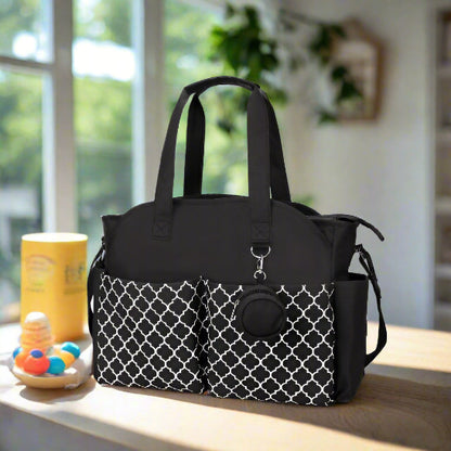 Le Sac pour Maternité est un sac noir de grande capacité avec deux poches avant présentant un design géométrique blanc, une fermeture éclair sur le dessus, des poignées noires, une bandoulière amovible et un accessoire rond sur le devant.