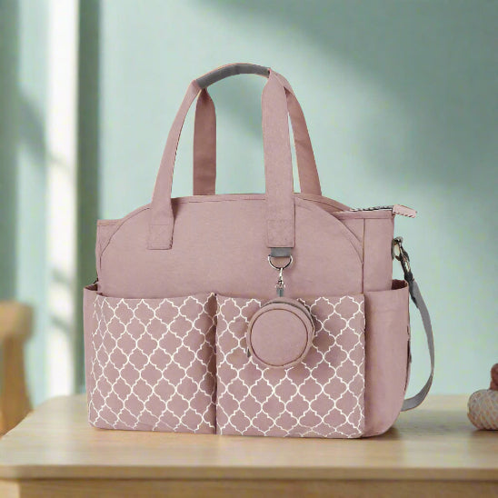 Un sac pour maternité rose clair avec une grande capacité, deux poches avant à motifs, des poignées supérieures, une bandoulière amovible et une pochette avant ronde est présenté vide sur un fond blanc.