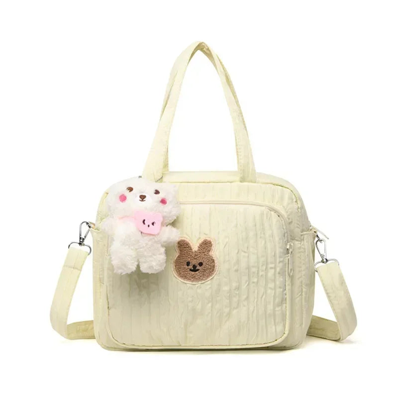 Le Sac pour la maternité | longchamp est un sac de maternité de couleur crème avec une poche zippée sur le devant, un petit patch lapin marron, un porte-clés en peluche cochon blanc, deux poignées et une bandoulière amovible, parfait pour les parents en déplacement.