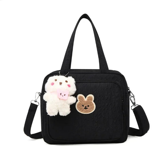 Le Sac pour la maternité | Longchamp est un sac noir avec une poche avant, orné d'un patch lapin marron et d'un porte-clés ourson en peluche blanche. Idéal pour les parents nomades, il possède deux anses et une bandoulière amovible.