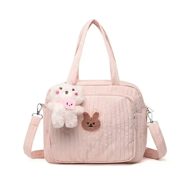 Le Sac pour la maternité | longchamp est un sac matelassé rose clair avec deux anses, une bandoulière amovible, un patch lapin marron sur la poche avant et un porte-clés animal en peluche, idéal pour les parents en déplacement.