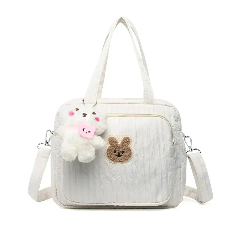 Le Sac pour la maternité | longchamp est un sac à langer matelassé blanc avec des bretelles, une longue bandoulière amovible, une poche avant avec un visage de lapin marron et un porte-clés ours en peluche avec un nœud rose, idéal pour les parents en déplacement.